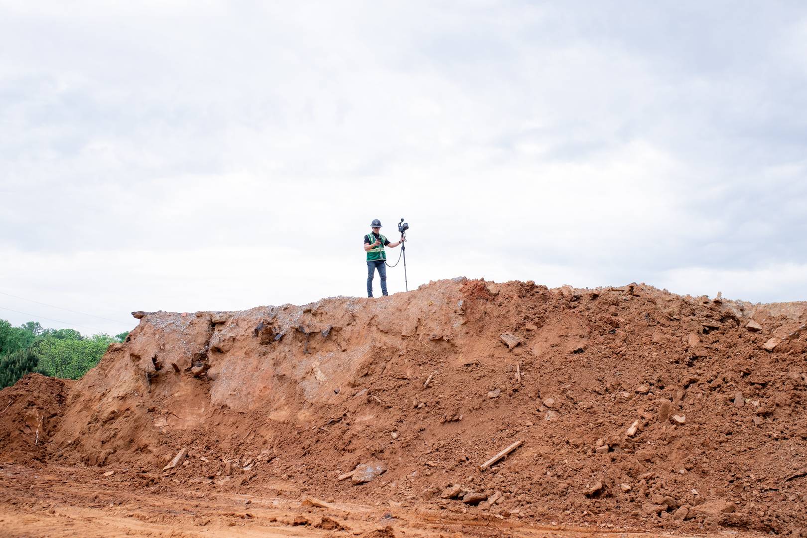 A person land surveying on top of a mud mound wearing personal protection equipment and holding a mobile 3d laser scanner