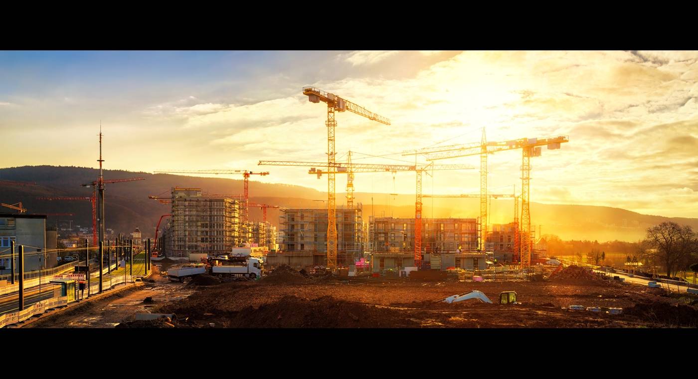 Panoramic view of a construction site at sunset, featuring multiple cranes, partially constructed buildings, and surrounding natural landscapes.
